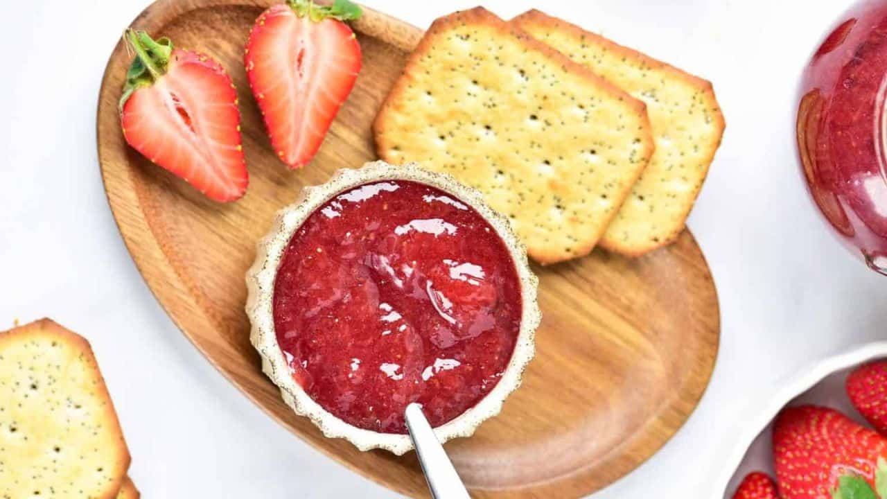 A small bowl of strawberry jam on a wooden tray with crackers and fresh strawberries.
