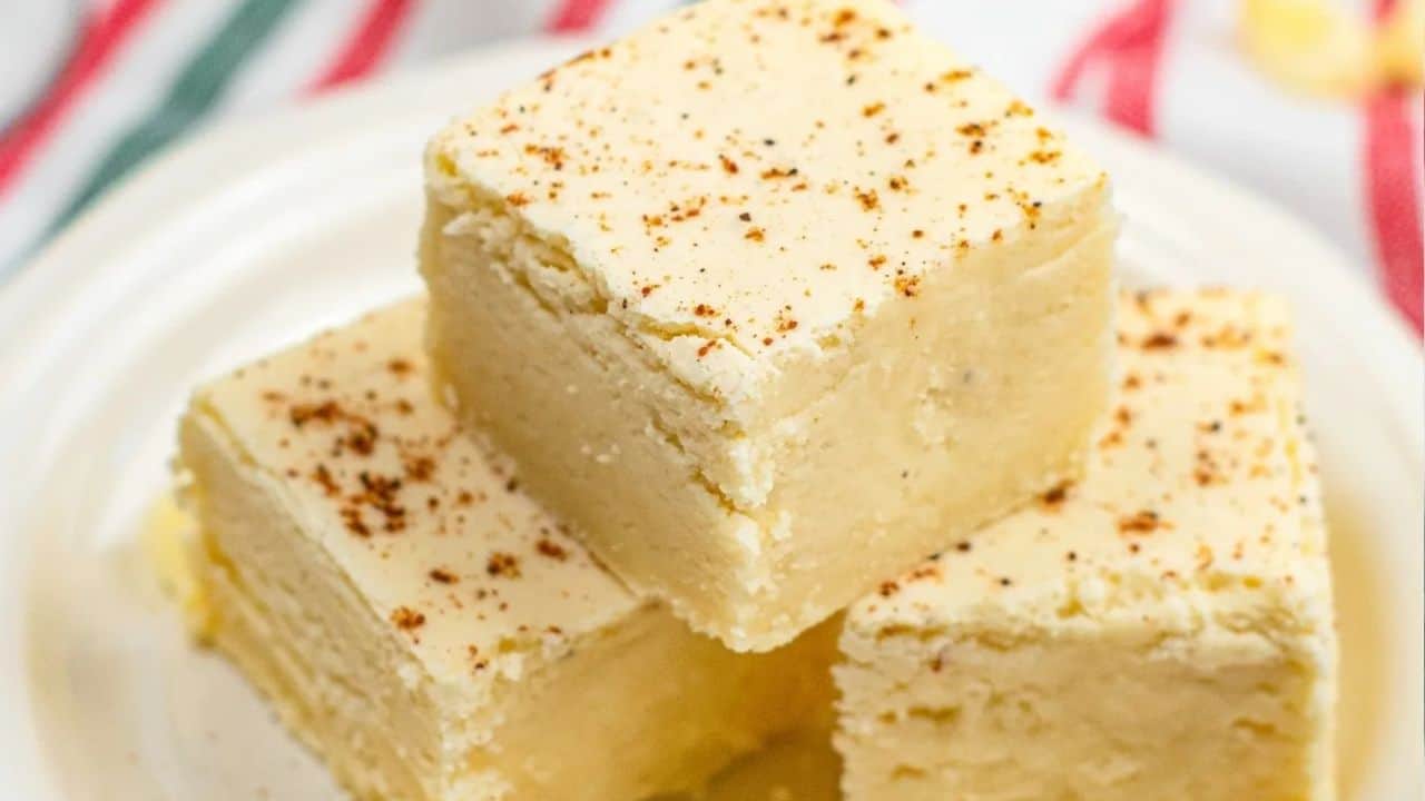 Three square pieces of pale yellow fudge, sprinkled with nutmeg, stacked on a white plate with a striped cloth in the background.