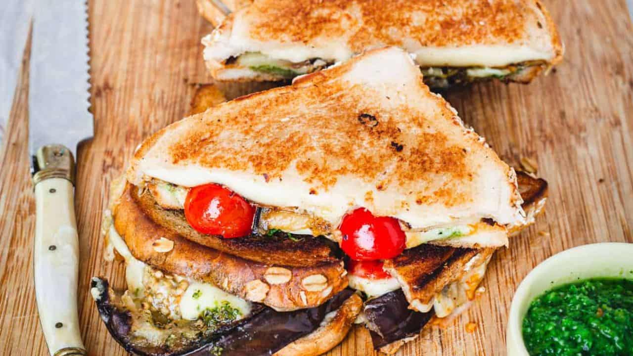 A grilled sandwich with mushrooms, tomatoes and pesto on a cutting board.