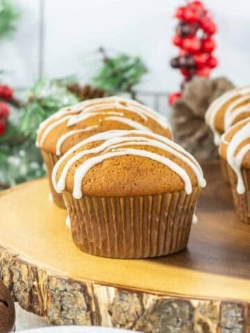 Gingerbread muffins with white icing drizzle are displayed on a wooden platter, with festive greenery and red berry decorations in the background.