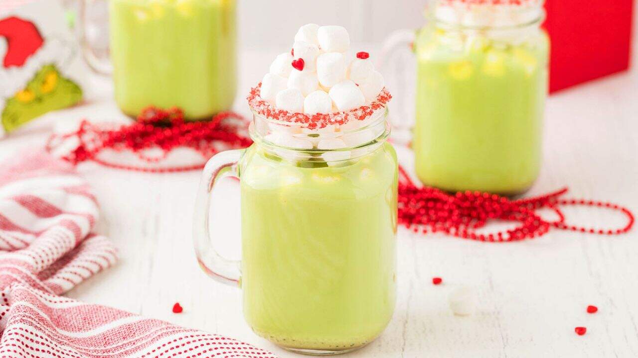 Three mason jars filled with green beverage, topped with mini marshmallows and red sprinkles, sit on a white table with red beads and a striped cloth nearby.