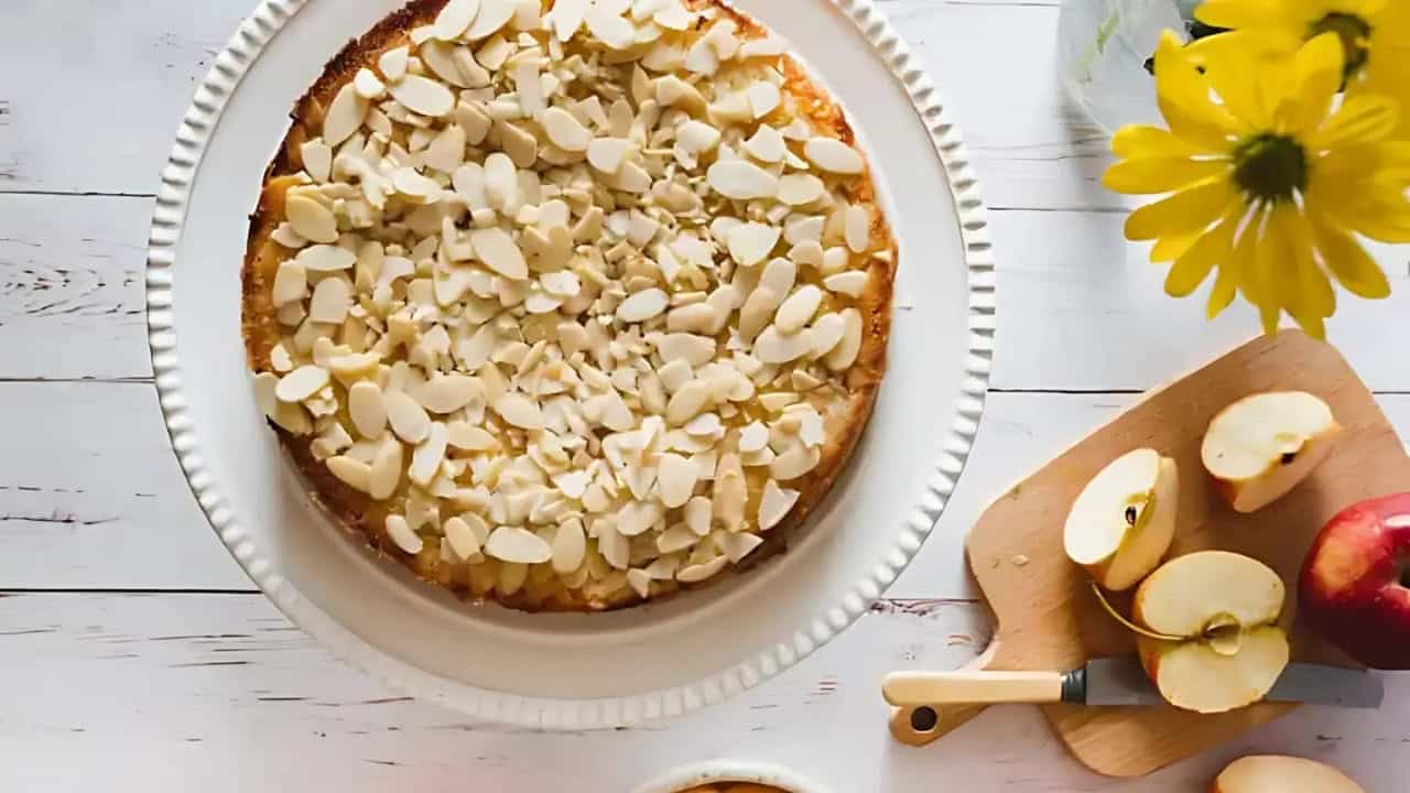 A round almond-topped cake on a white plate next to a cutting board with sliced apples, a knife, yellow flowers, and a cup on a white wooden surface.