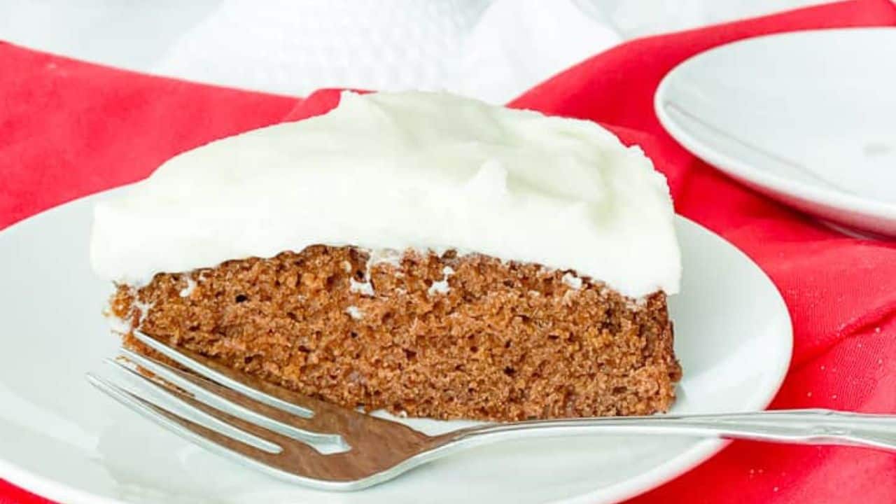 A slice of gingerbread cake with creamy frosting on a white plate, next to the rest of the cake.
