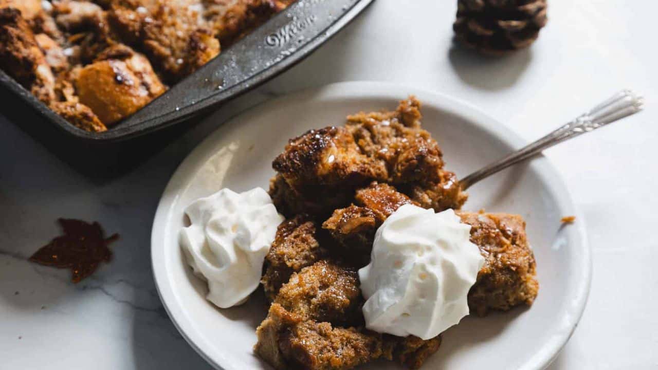 A plate of bread pudding topped with two dollops of whipped cream. A fork rests on the plate. In the background, a baking dish with more bread pudding is partially visible on a marble surface. Decorative items are placed around.