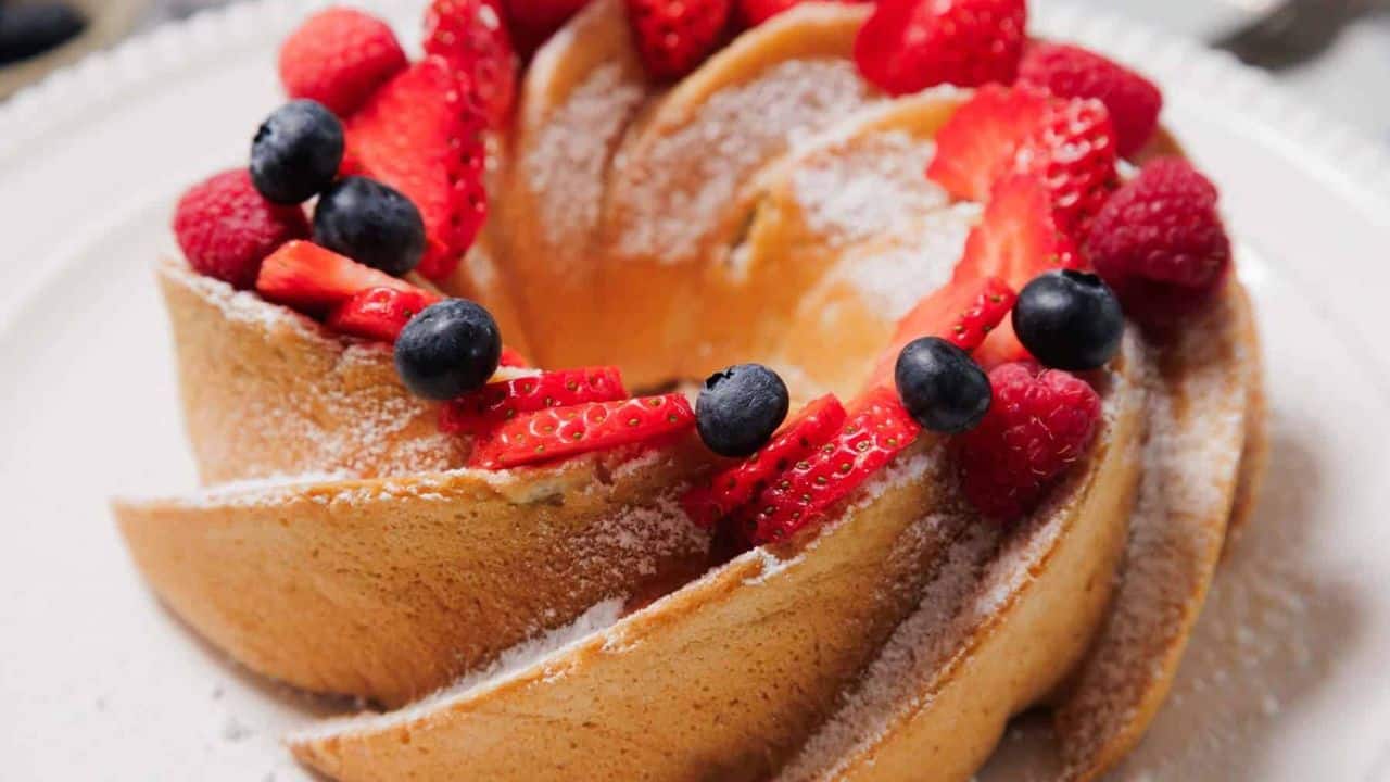 A Bundt cake topped with sliced strawberries, whole blueberries, and a dusting of powdered sugar sits on a white plate. A bowl of blueberries and a decorative plate are visible in the background.