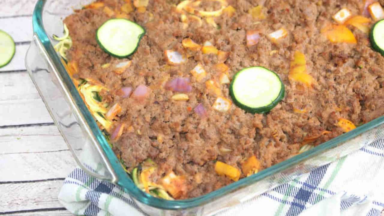 A glass baking dish filled with a ground meat and vegetable casserole, topped with zucchini slices and diced yellow bell peppers on a wooden surface.