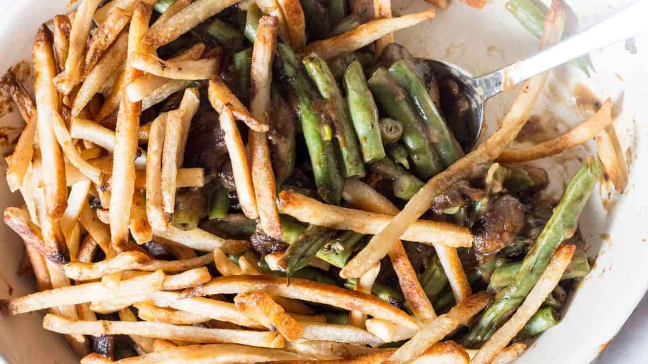 A close-up of a dish with green beans, thin French fries, and mushrooms, mixed together with a spoon.