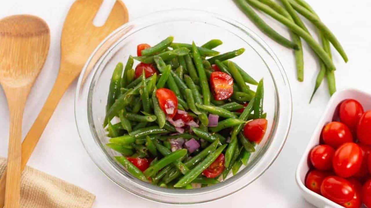 A plate of green beans tossed with cherry tomatoes and crumbled feta cheese, accompanied by a fork, on a white surface.