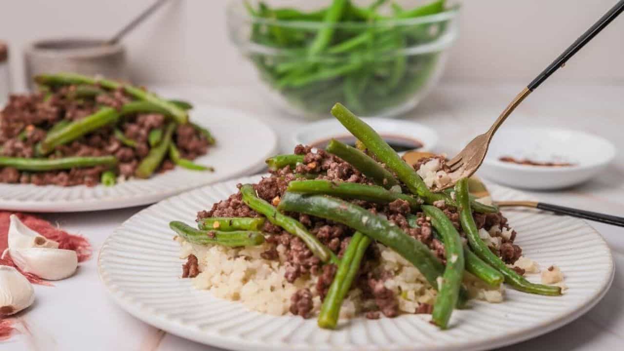 Plate with ground beef, green beans, and cauliflower rice. Fork on the plate, garlic cloves nearby, and a bowl of green beans in the background.