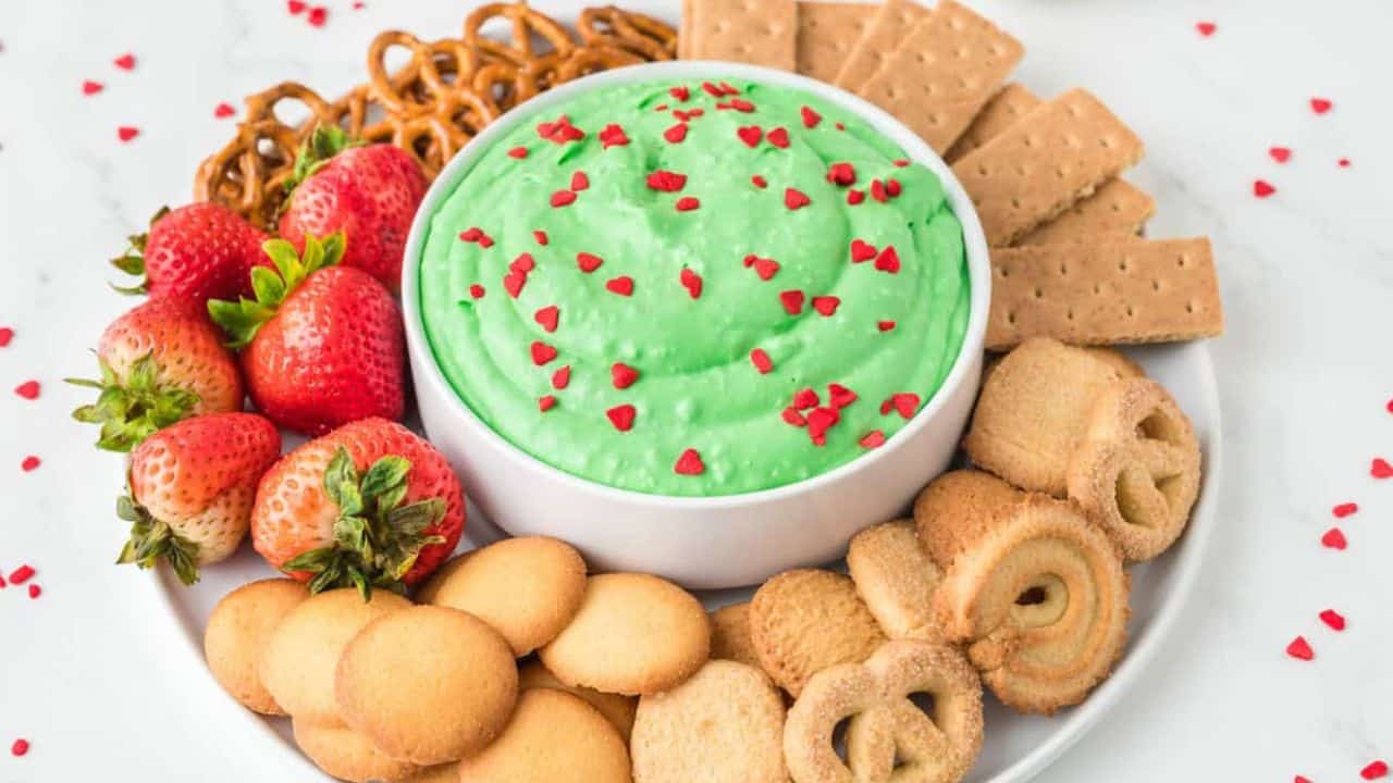 A close up image of grinch dip in the center with an assortment of crackers, biscuits, pretzels, and strawberries on the side.