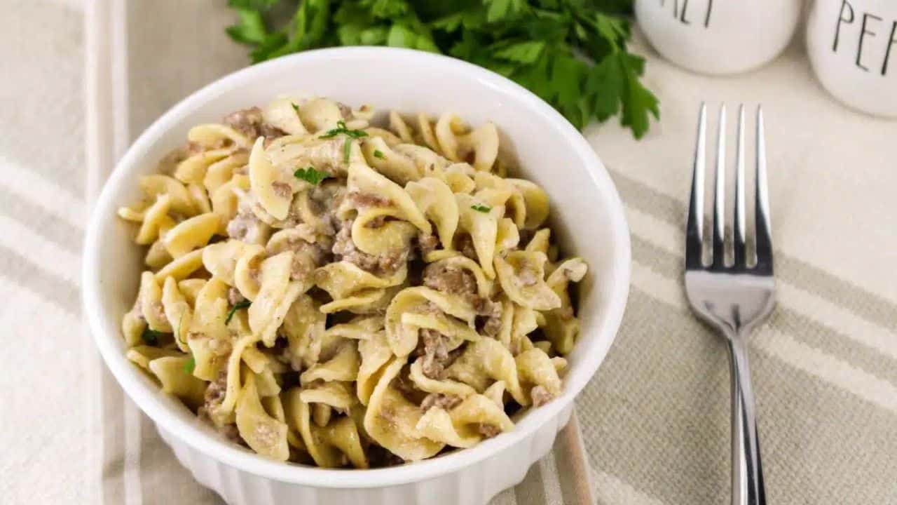 A bowl of creamy beef stroganoff with egg noodles on a striped cloth next to a fork and a sprig of parsley.