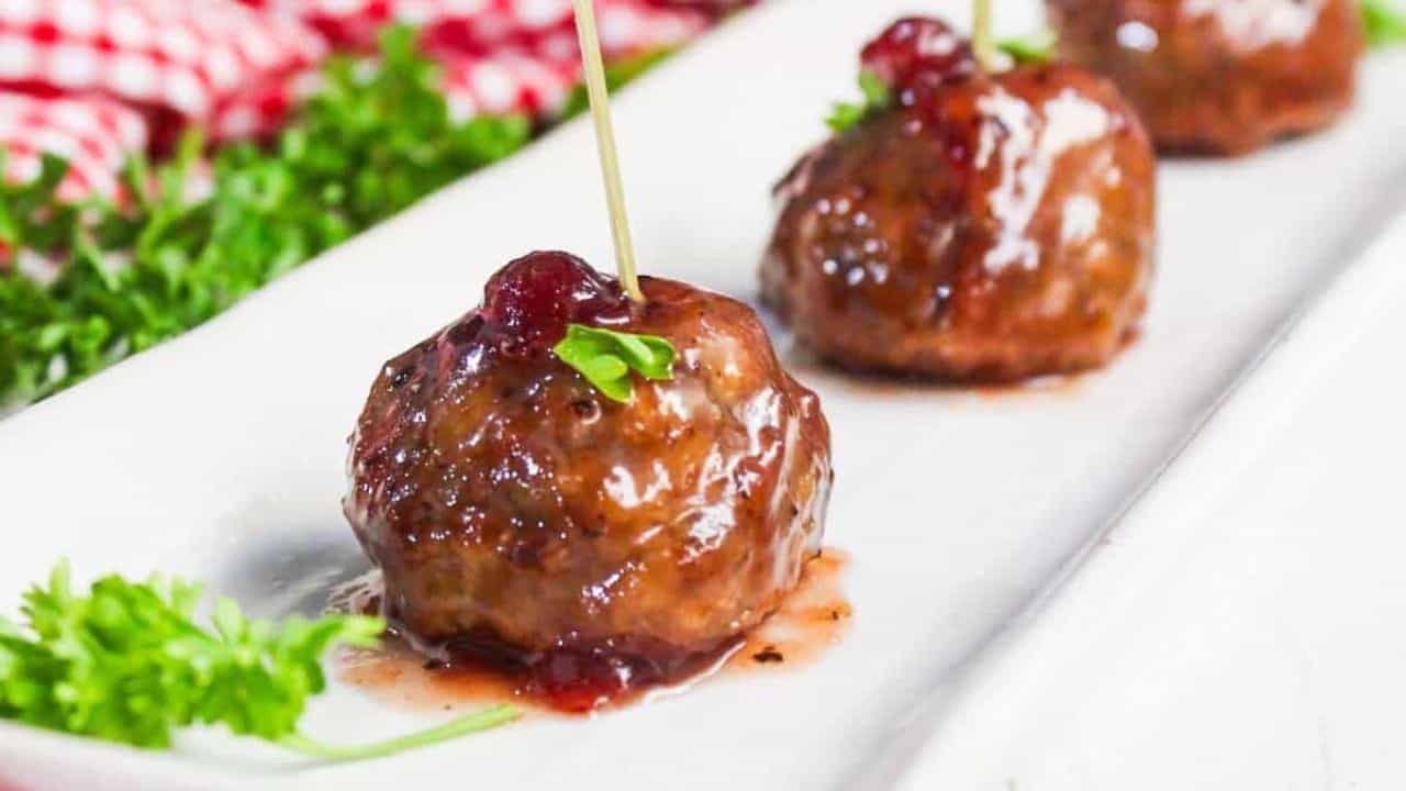 Three glazed meatballs are arranged in a row on a white plate, each garnished with a stick and a small sprig of parsley. A red checkered cloth and some fresh parsley are in the background.