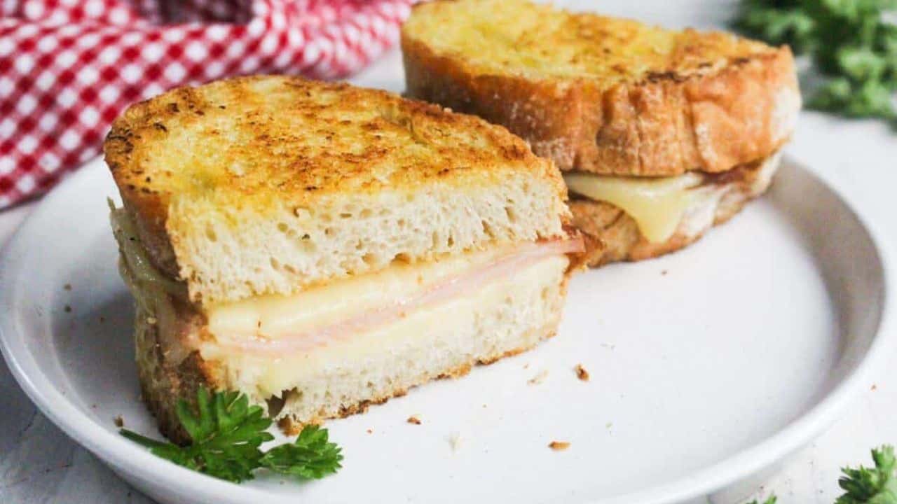 A grilled cheese sandwich with ham is halved and placed on a white plate. A red checkered cloth and green herbs are in the background.