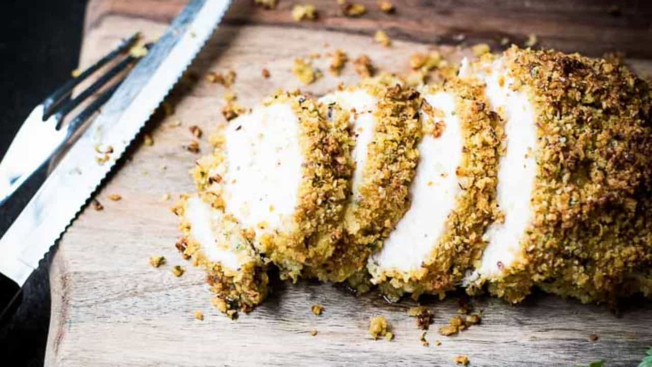 Sliced breaded chicken on a wooden cutting board next to a serrated knife.