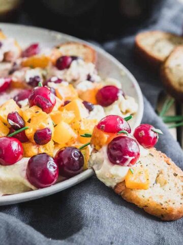 Crostini topped with creamy cheese, diced yellow fruit, cranberries, and herbs on a plate with more crostini on the side.
