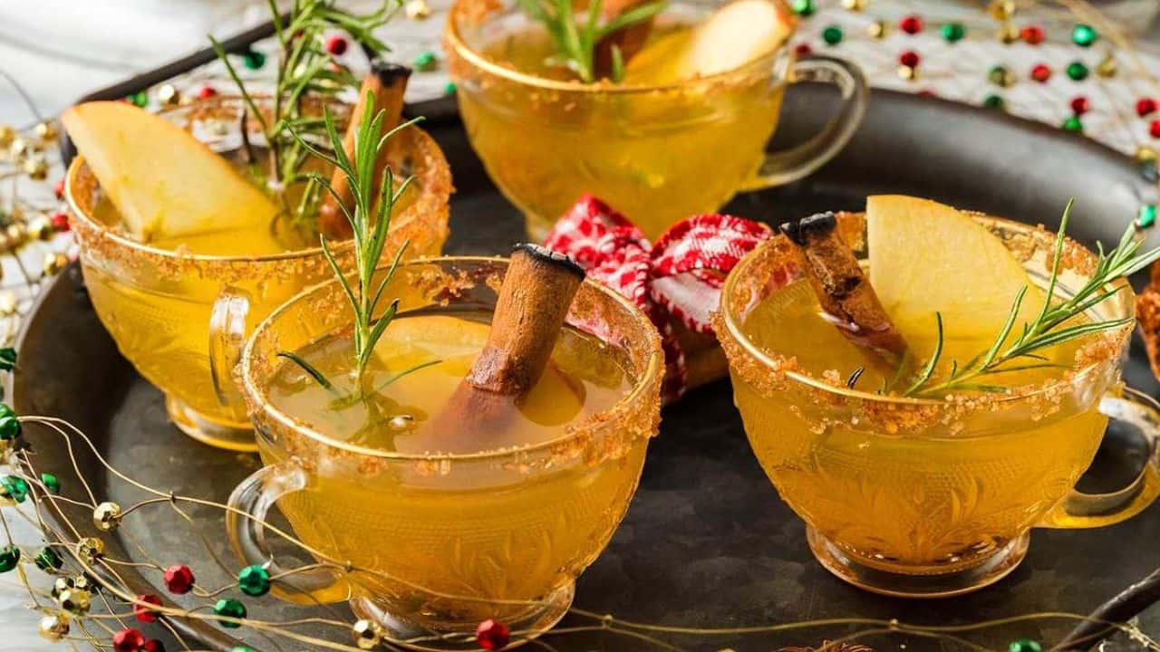 Four glass cups of yellow beverage garnished with apple slices, rosemary sprigs, and cinnamon sticks are arranged on a tray with festive decorations.