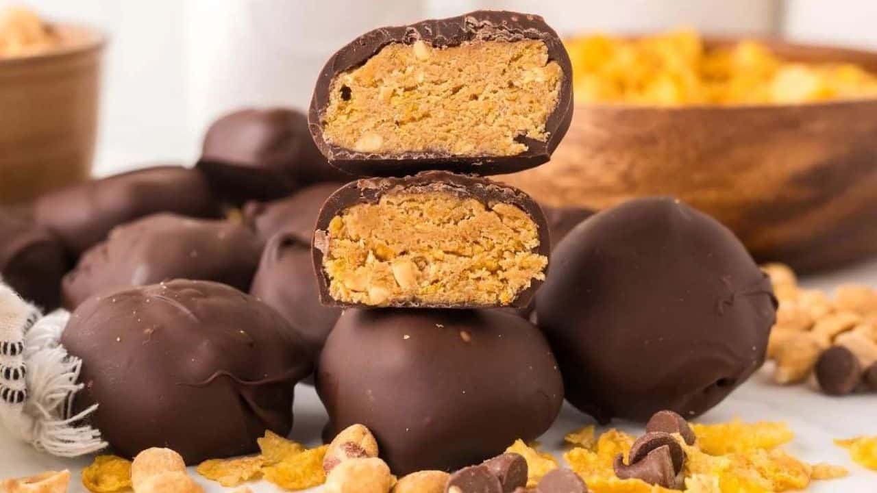 Chocolate-covered peanut butter balls are stacked, with one cut in half to show the crumbly peanut butter filling inside. Scattered peanuts, chocolate chips, and cereal are visible around them.