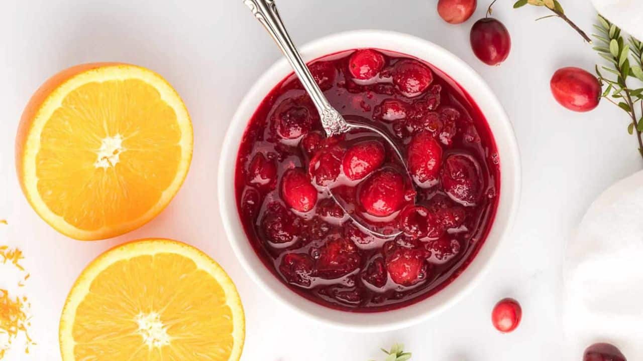 A close up photo of fresh cranberry sauce placed in a white bowl with spoon, oranges and fresh cranberries in the background.