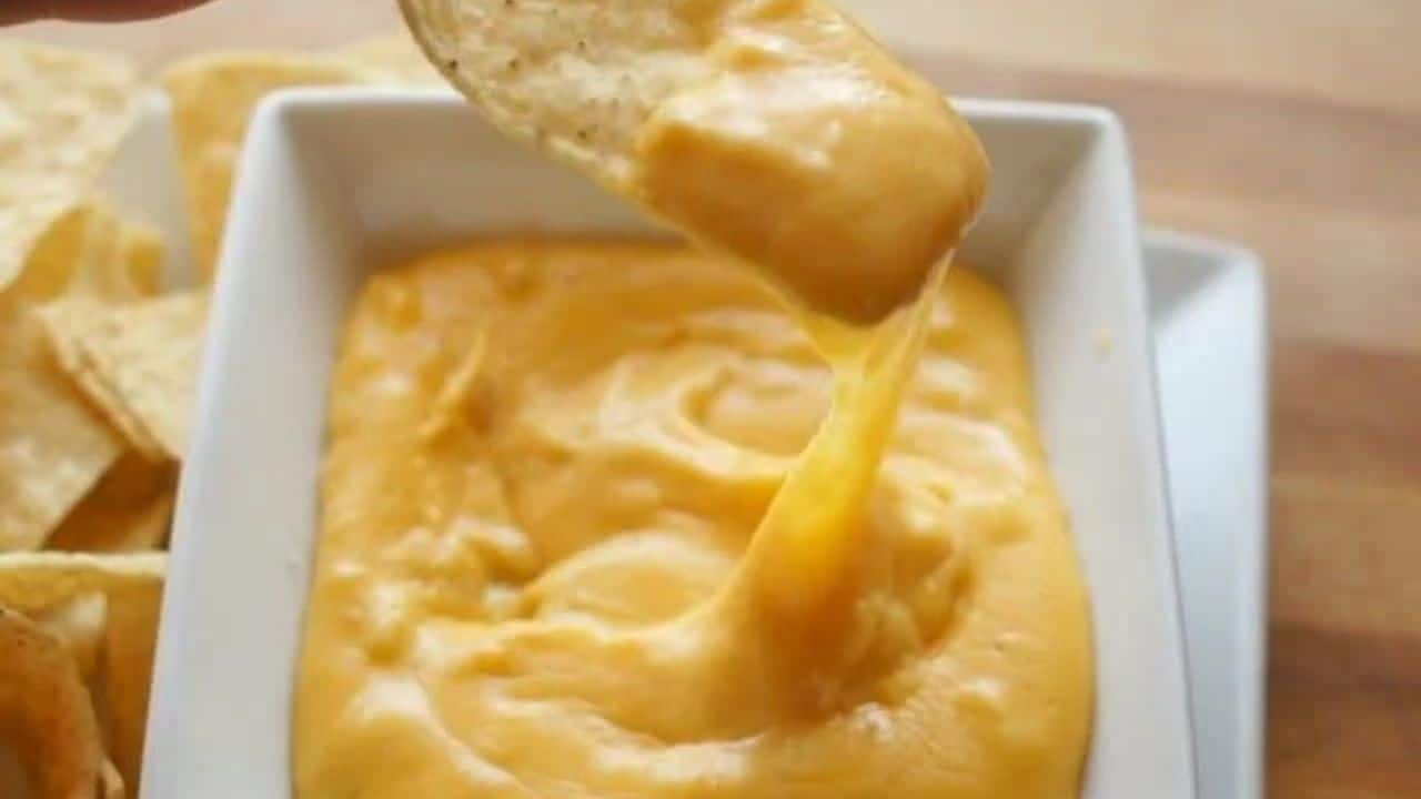 Image shows a tortilla chip pulling nacho cheese out of a white bowl filled with it.