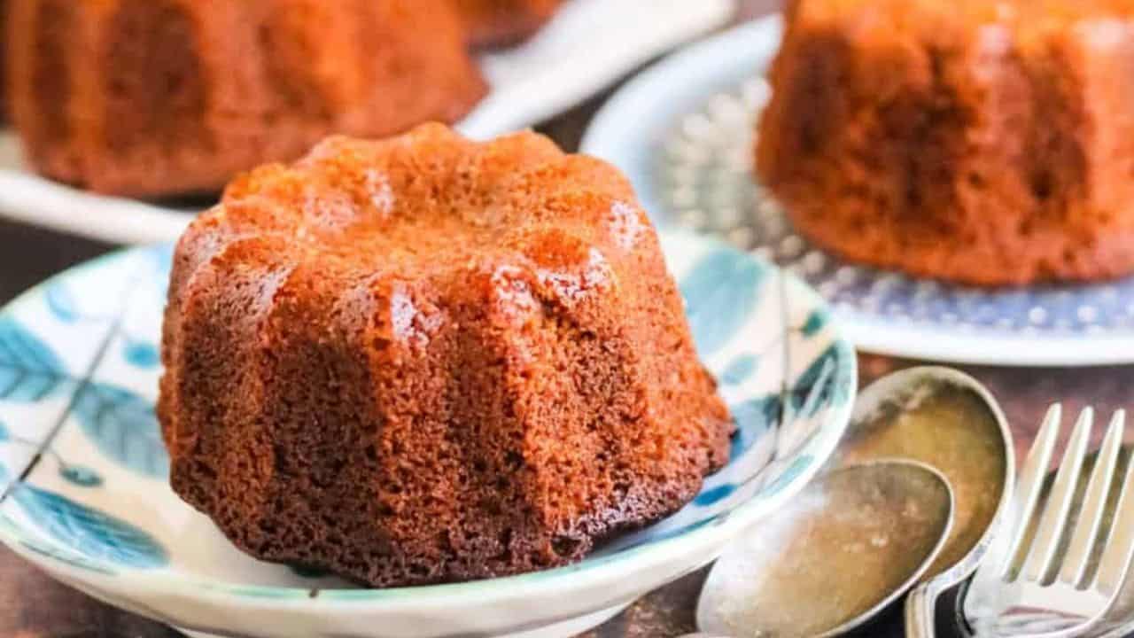 A small, brown, bundt-shaped cake is served on a decorative plate with spoons and a fork beside it; more cakes are visible in the background.