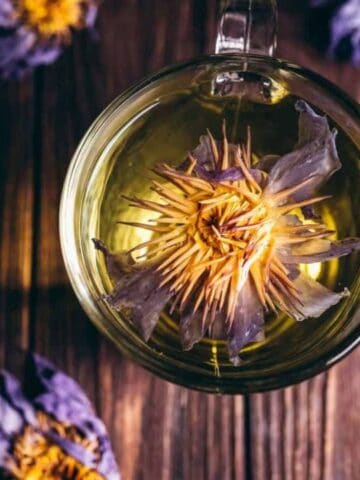A glass cup of tea with a blooming purple flower inside, viewed from above, on a wooden surface with dried flowers scattered around.