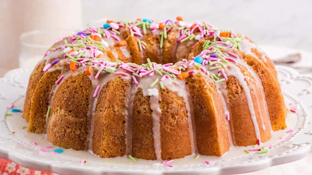 A Bundt cake with white icing drips and colorful sprinkles sits on a white decorative cake stand.
