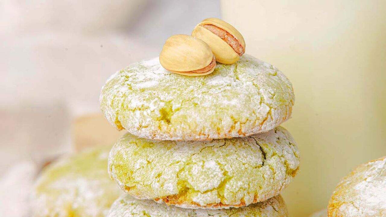 Three pistachio cookies dusted with powdered sugar are stacked on top of each other, with two whole pistachios placed on the top cookie.