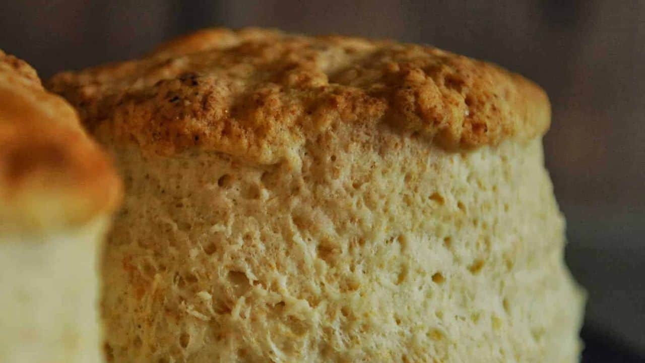 Close-up of a freshly baked, golden-brown scone with a textured, flaky surface reminiscent of cream biscuits on a dark background.