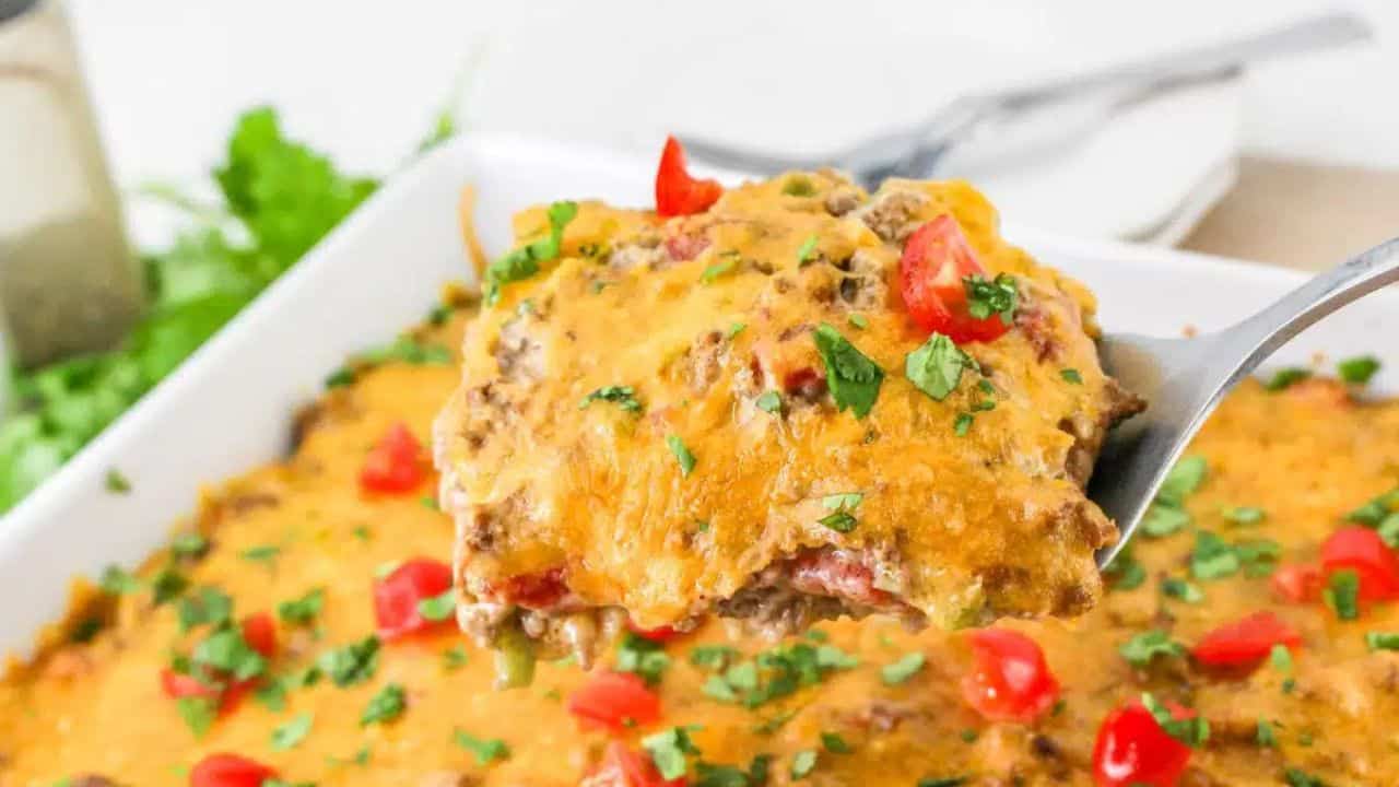 Close-up of a spoon lifting a portion of cheesy beef casserole garnished with chopped tomatoes and herbs from a white baking dish.