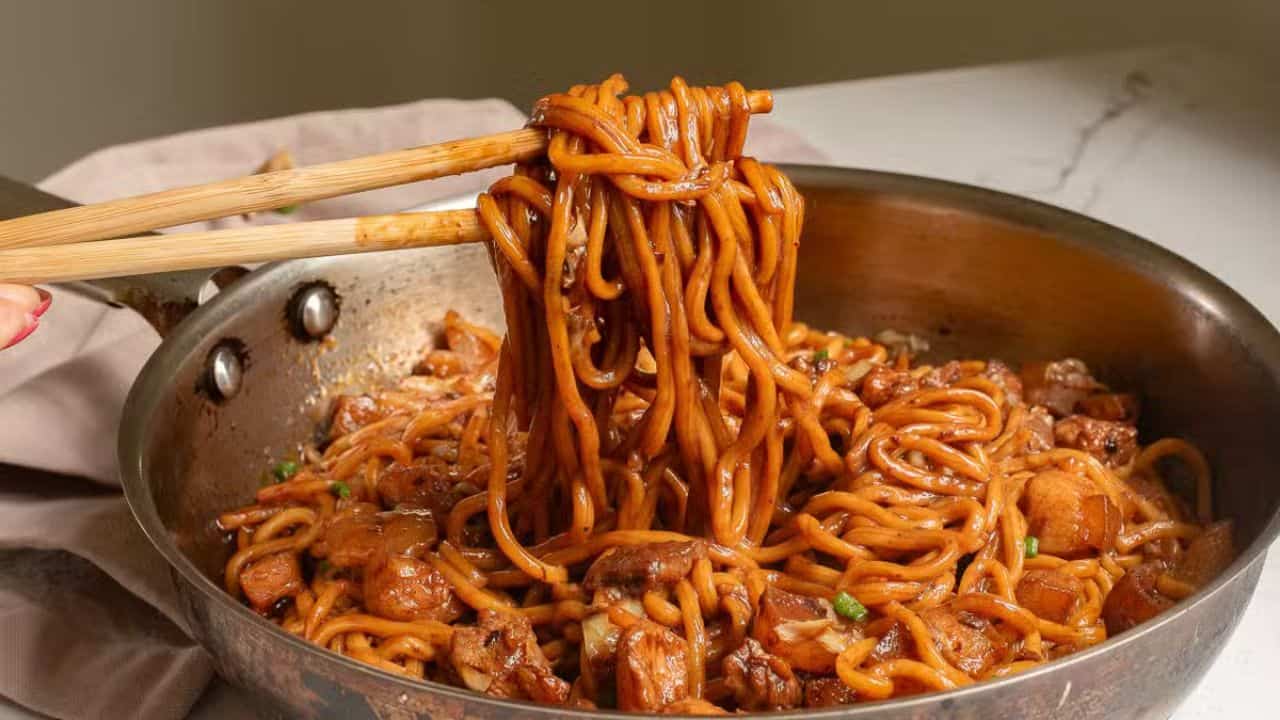 A close-up of a pan filled with savory noodles and chunks of meat being lifted by chopsticks. The dish appears well-seasoned with sauce, and green garnishes are sprinkled on top. A beige napkin and a piece of dumpling are in the background.