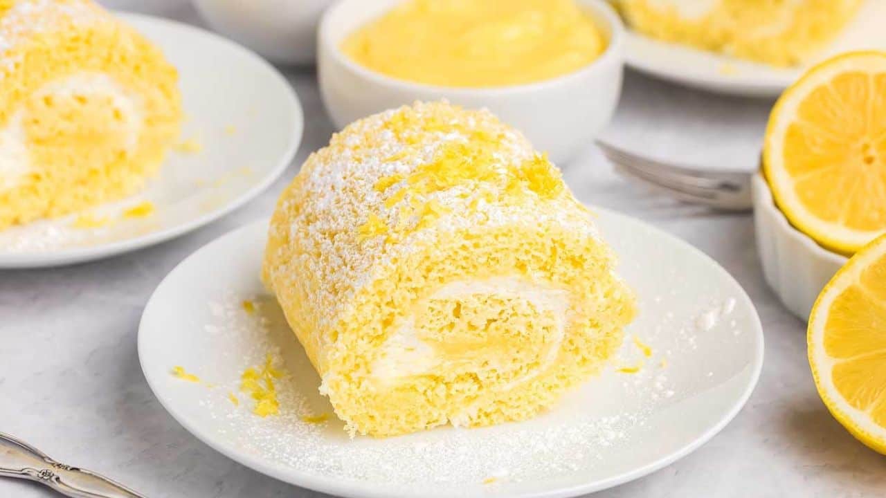 A slice of lemon Swiss roll cake with powdered sugar on top sits on a white plate, with lemons and a bowl of lemon filling in the background.