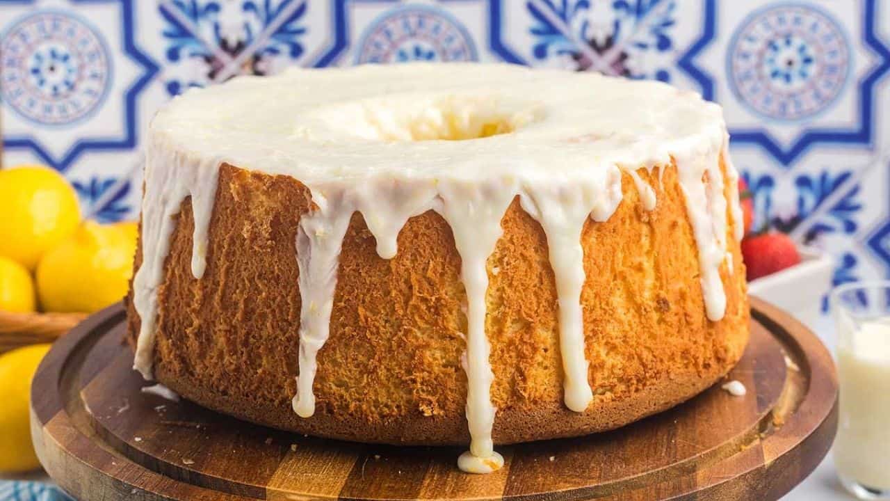 A round, golden bundt cake with white icing drips sits on a wooden board, with lemons, strawberries, and a blue patterned tile background.