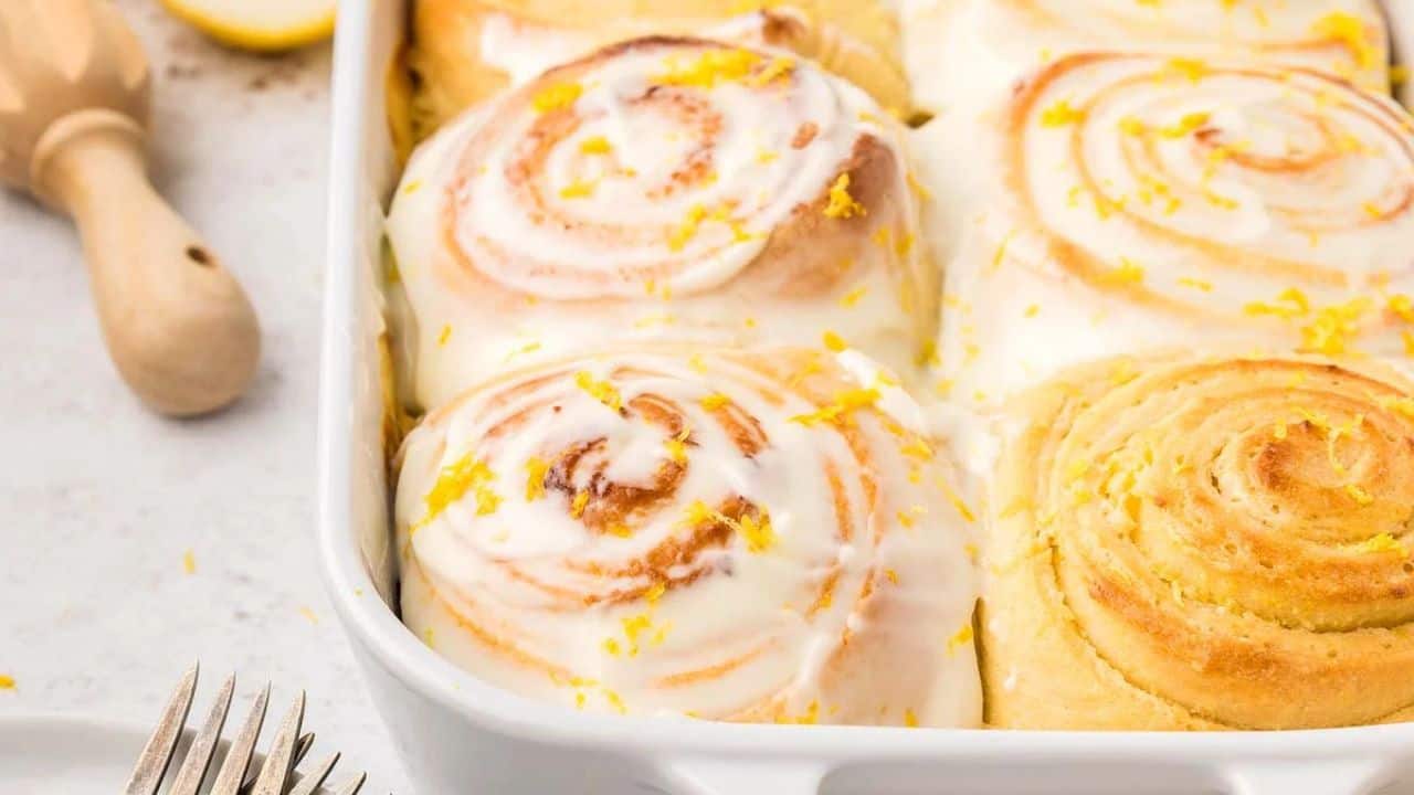 A baking dish filled with iced cinnamon rolls topped with lemon zest, with a wooden citrus reamer and lemon wedge nearby.