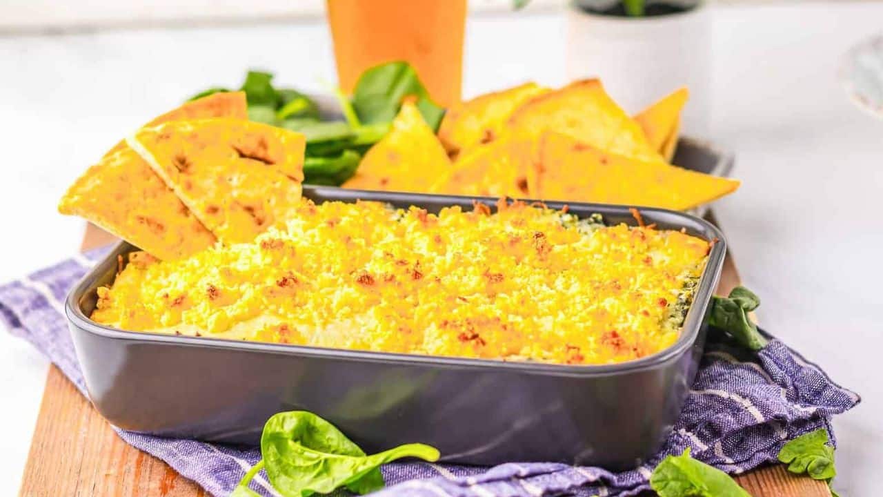 A close-up of spinach artichoke dip on a cracker held by a hand. The dip is creamy with visible green spinach and bits of artichoke. The background shows a baking dish filled with the dip.