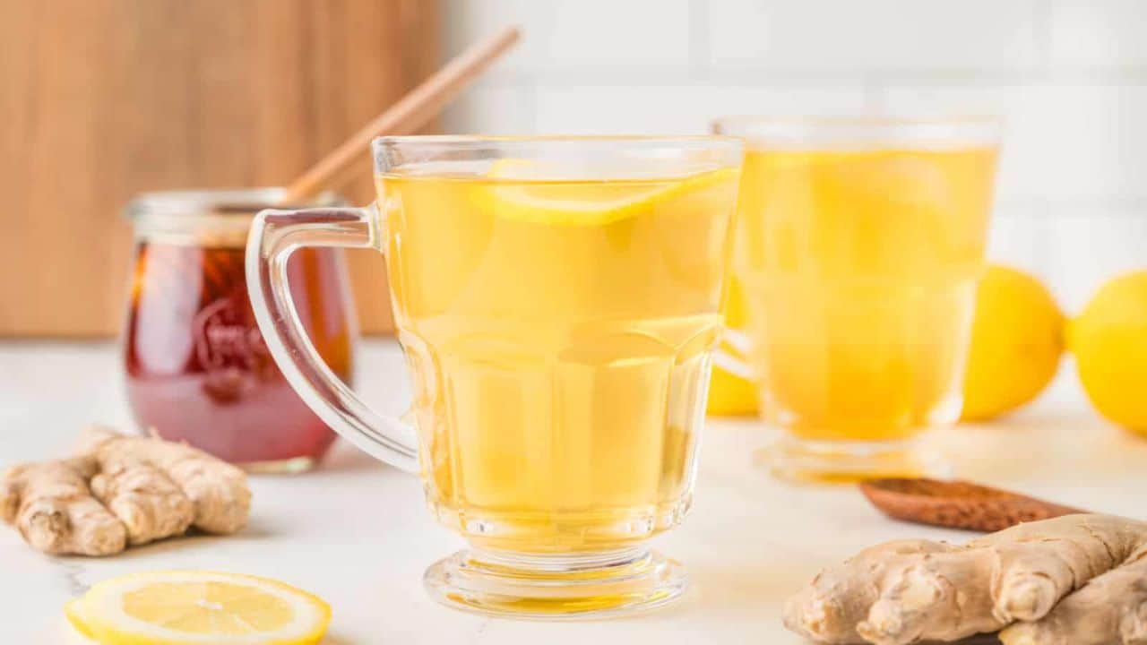White cup of ginger tea on a saucer, with ginger root, sliced lemon, honey with a dipper, and a wooden spoon on a light surface.