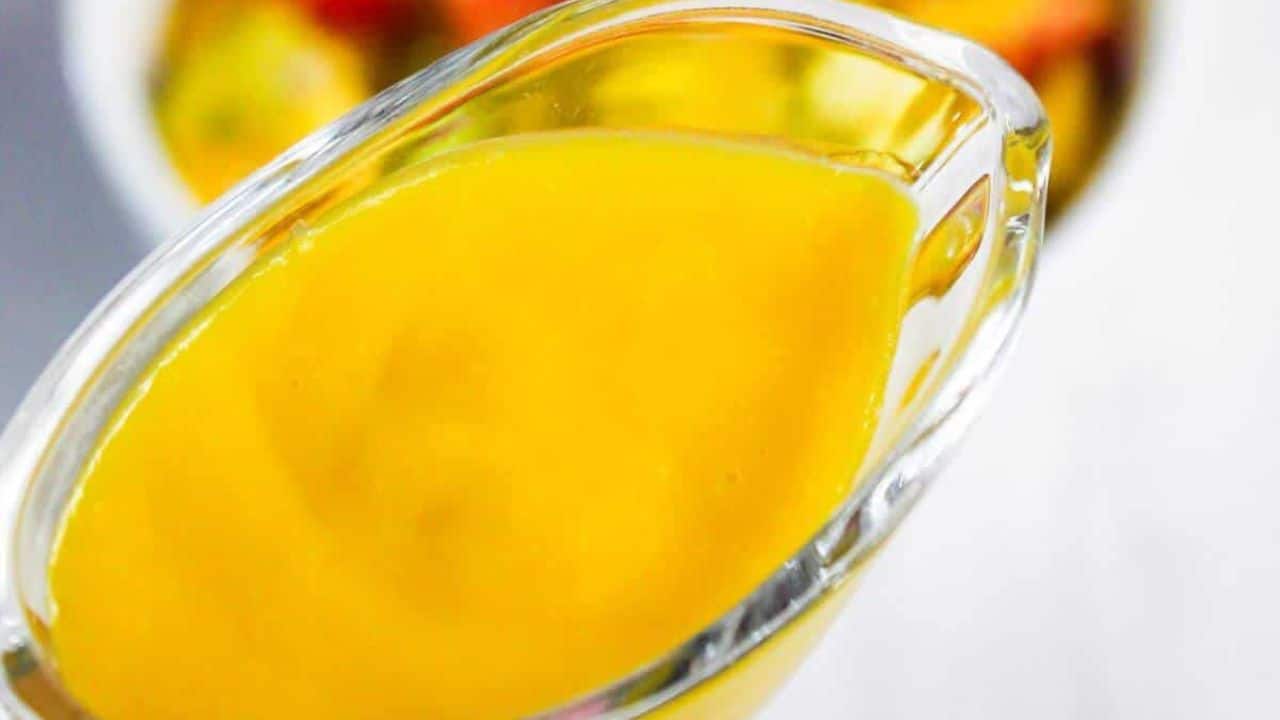 A glass container pours yellow sauce over grilled shrimp in a bowl with mango slices, nuts, and leafy greens.