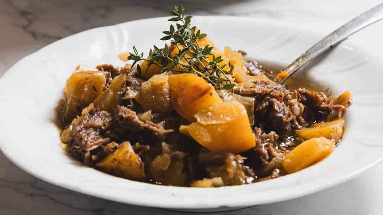 A white plate containing a stew made of tender shredded meat and chunks of potatoes, garnished with sprigs of fresh thyme. A spoon rests on the side of the dish, placed on a marble surface.