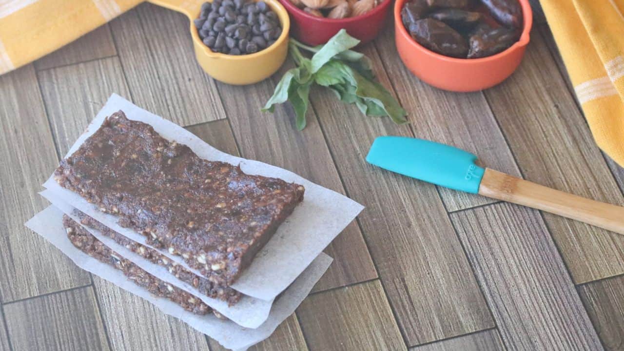 Three chocolate protein bars stacked with parchment paper, next to bowls of chocolate chips, almonds, dates, and a spatula on a wooden surface.