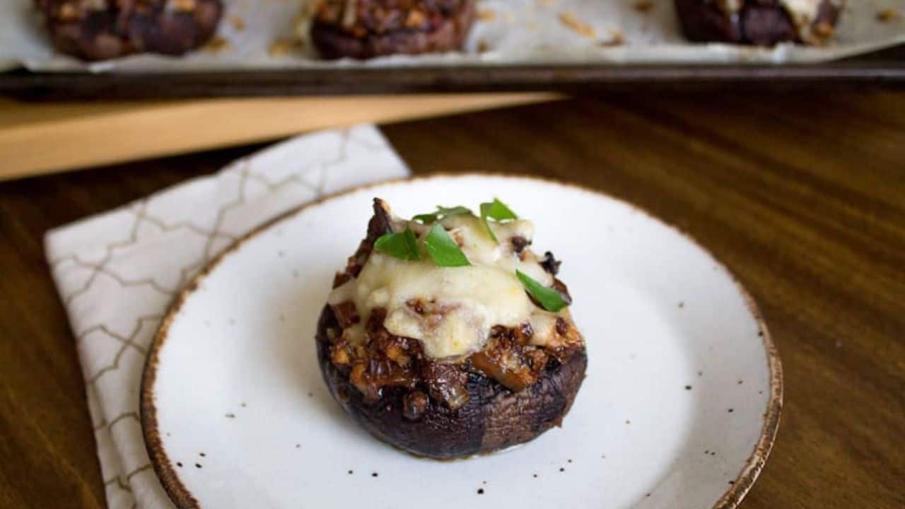 A stuffed mushroom topped with melted cheese and herbs sits on a white plate with a brown rim; more stuffed mushrooms are on a tray in the background.