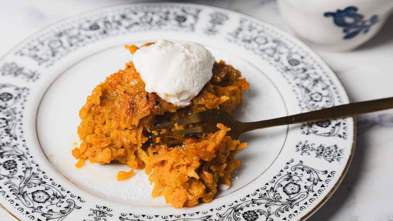 A delightful slice of carrot cake with a dollop of whipped cream graces a decorative black and white plate. A fork rests invitingly on the cake while a white and blue cup peeks from the background, almost like the perfect ending to a recipe for an unforgettable carrot casserole.