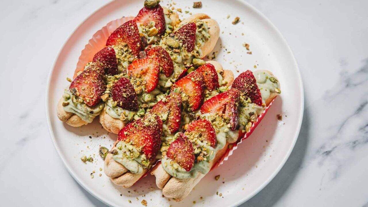 Three ladyfinger desserts topped with green cream, sliced strawberries, and chopped pistachios on a white plate.