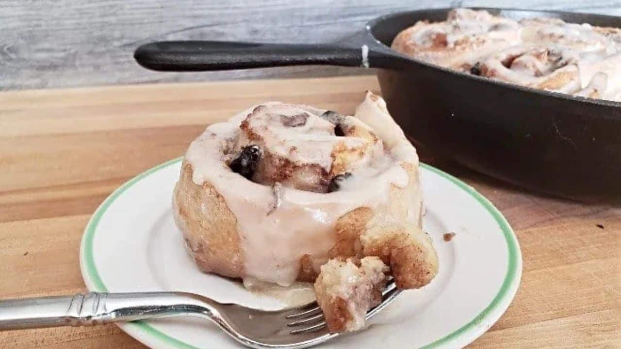 Image shows a cinnamon roll on a plate with a fork holding a bite and the full pan behind it.