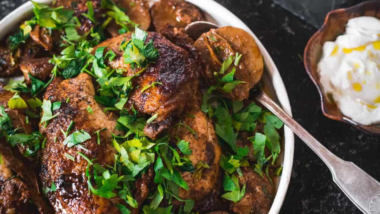 A plate of roasted chicken with crispy skin, garnished with chopped green herbs. Beside the chicken are sliced potatoes and mushrooms. An ornate fork is placed on the plate, which has a decorative floral pattern.