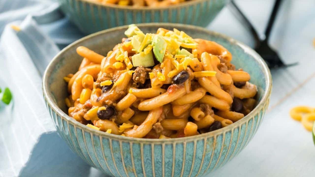 A bowl of macaroni mixed with ground meat, black beans, and topped with shredded cheese and diced avocado.