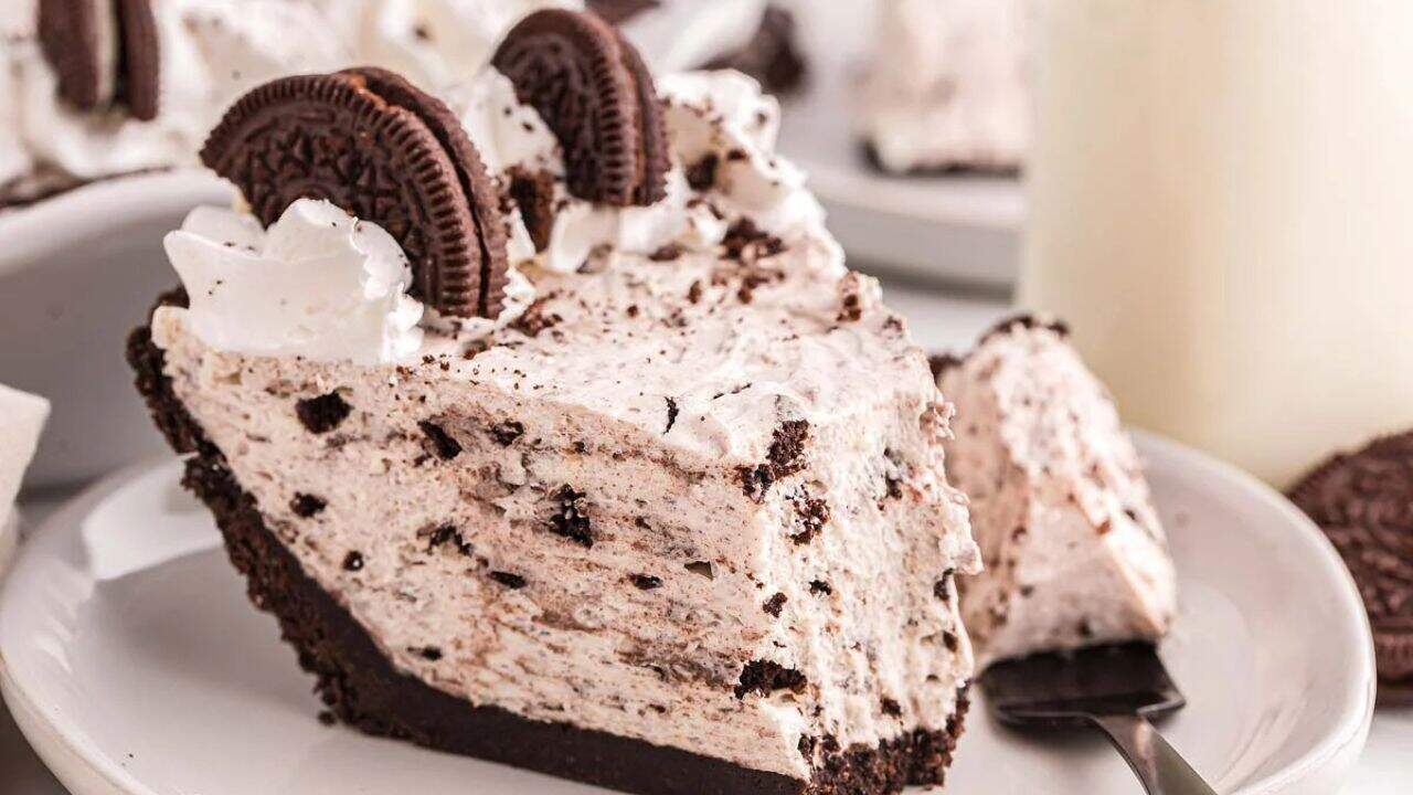 A slice of cookies and cream pie with a chocolate cookie crust, topped with whipped cream and two chocolate sandwich cookies on a white plate.