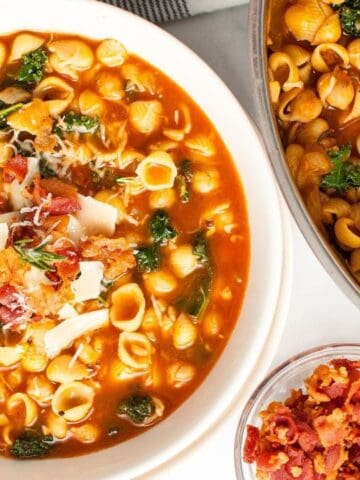 A bowl and pot of shell pasta in a tomato-based broth with greens, topped with grated cheese and bacon; a small bowl of bacon bits is nearby.