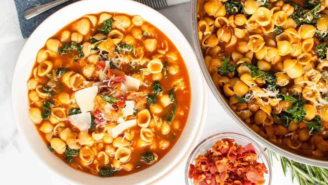 A bowl and pot of shell pasta in a tomato-based broth with greens, topped with grated cheese and bacon; a small bowl of bacon bits is nearby.