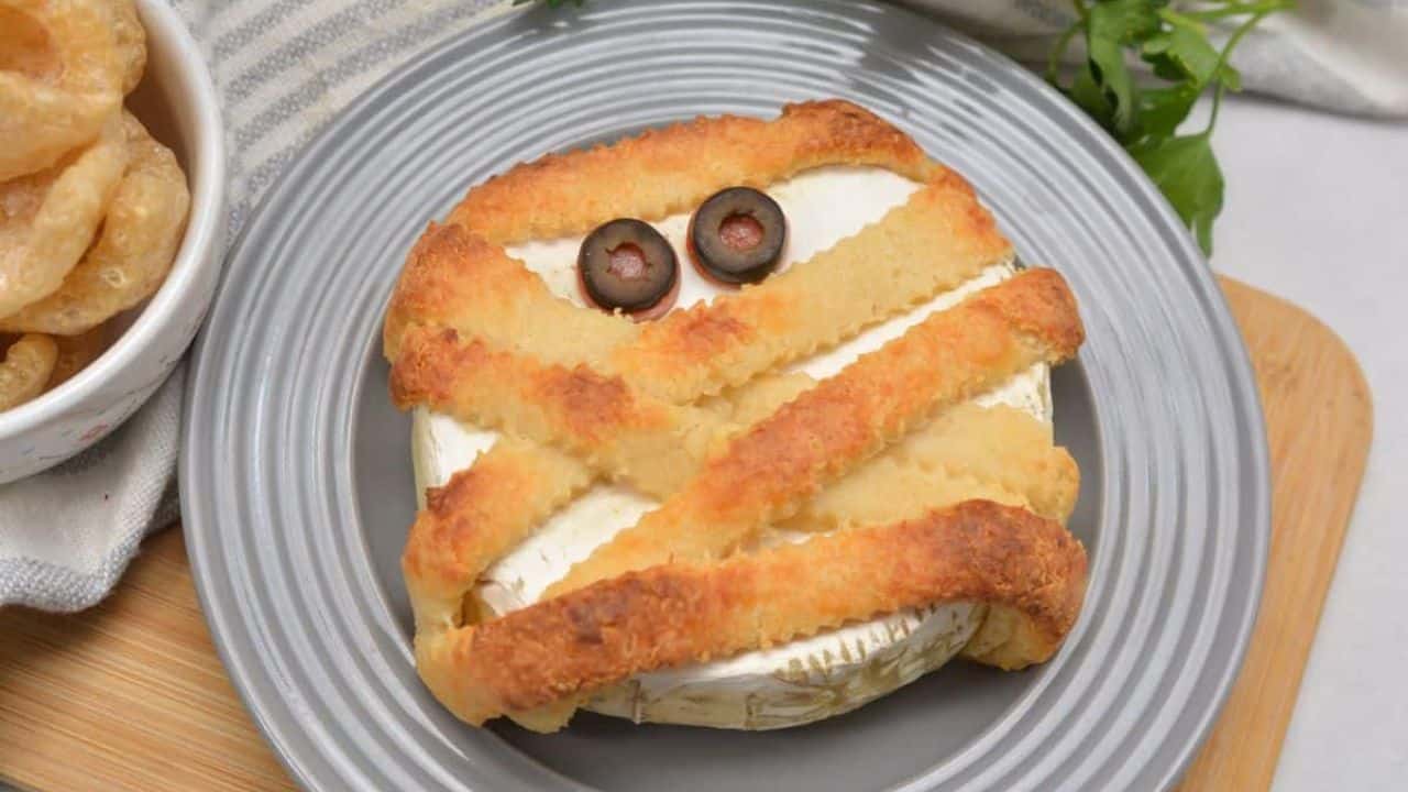 A mummy brie cheese with olives and pork rinds on a plate.