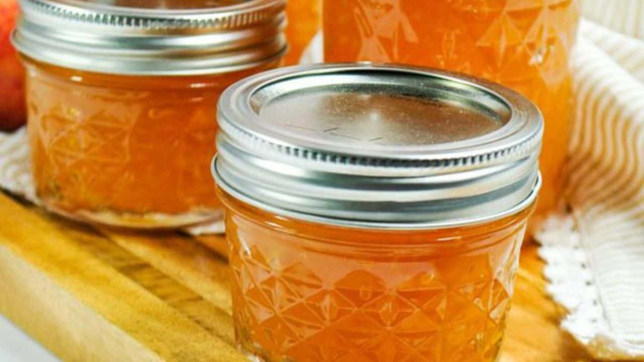 Three glass jars filled with orange-colored jam sit on a wooden surface, each sealed with a metal lid.