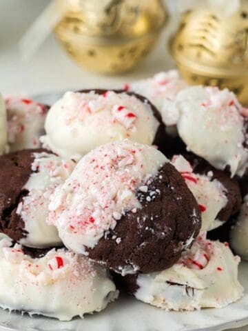 A plate of chocolate cookies partially coated in white icing and topped with crushed peppermint candy, with festive ornaments and candies in the background.